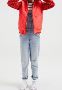 Czerwona pikowana kurtka bomberka z wzorowanymi rękawami, noszona na szarych dżinsowych ogrodniczkach. Białe trampki i w paski koszulka dopełniają stylizację.