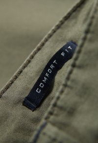 Grön tyg med en slät textur, med en svart etikett som har texten "COMFORT FIT" i vitt. Synlig sömnad längs kanten.