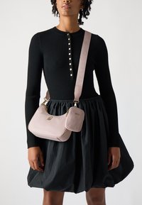 Piccola borsa a tracolla in pelle rosa con un logo dorato, chiusura a zip e tracolla staccabile. Indossata sopra un vestito lungo a maniche nere a coste con gonna ampia.
