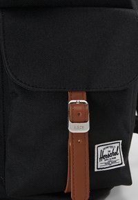 Zwarte stoffen rugzak met een voorklep, een bruin leren riem en een zilveren gesp. Voorzien van een wit merkzegel met blauw marineblauwe tekst.