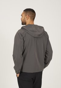 Grijze lichtgewicht jack met een capuchon, voorzien van elastische manchetten en een eenvoudig ontwerp, weergegeven van achteren tegen een effen witte achtergrond.