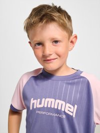 Junger Junge mit kurzen blonden Haaren, der ein lila-rosa Hummel Performance Sportshirt trägt und sanft vor einfarbigem Hintergrund lächelt.