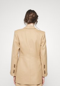 Veste blazer beige ajustée avec un design structuré, dotée d'un col cranté, de deux fentes arrière et de poignets avec boutons. Tissu à texture lisse.