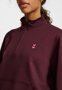 Burgundy kvart-zip hoodie med en mjuk textur, med en liten rosa logotyp och ficka på framsidan. Tyget verkar mjukt och avslappnat.