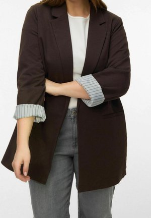 Personne portant un blazer marron foncé avec des poignets rayés retroussés, une chemise blanche et un jean gris sur un fond blanc uni.