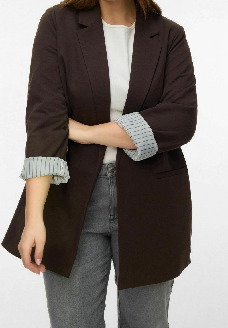 Personne portant un blazer marron foncé avec des poignets rayés retroussés, une chemise blanche et un jean gris sur un fond blanc uni.