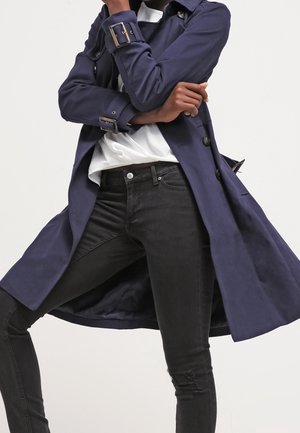 Osoba ubrana w granatowy płaszcz przeciwdeszczowy, na białej koszuli i czarnych dżinsach, stojąca złożonymi ramionami i jedną nogą zgiętą.