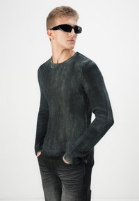Tamsiai žalia raukinėta megztukas su ilgomis rankovėmis, apvaliu iškirpimu, dėvimas su slim-fit tamsiomis džinsais ir juodais saulės akinius. Lygus paviršius.