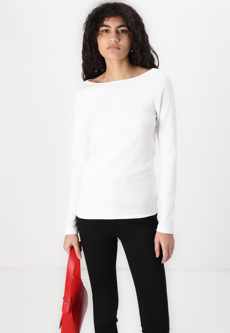 Weißes Langarmoberteil mit einem Bootsausschnitt, kombiniert mit schwarzen Hosen. Das Model hält eine rote Lederhandtasche. Sanfte Textur; minimalistisches Design.
