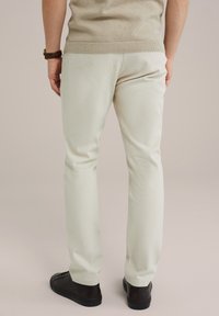Light beige trousers made of textured fabric, featuring a straight-leg cut and paired with dark brown sneakers. Seen from the back.
