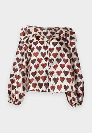 Frau trägt eine weiße Bluse mit roten Herzmustern, beige weite Hosen und rote Schuhe, stehend vor einem einfarbigen hellgrauen Hintergrund.