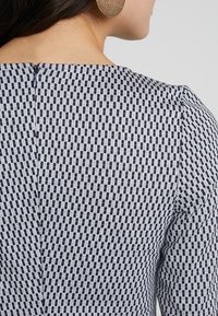 Námořnické a bílé šaty se vzorem z texturované látky, s bočním zipem a přiléhavým střihem, zdůrazňující geometrické tvary.