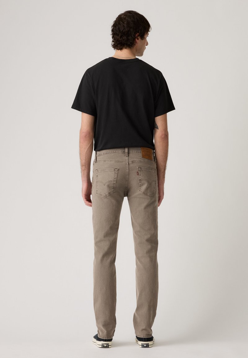 Uomo con maglietta nera, jeans beige e sneakers nere, in piedi di spalle contro uno sfondo chiaro e uniforme.