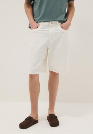 Persona che indossa pantaloncini bianchi fino al ginocchio, una maglietta turchese e scarpe slip-on marroni, in piedi con le mani nelle tasche davanti a uno sfondo semplice.