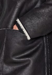 Main partiellement dans la poche d’un manteau texturé foncé avec une doublure en fourrure blanche visible au niveau du poignet.