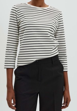 Personne portant une chemise blanche à manches longues avec des rayures horizontales noires et un pantalon noir sur fond clair uni.