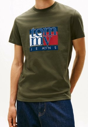 Mand iført en olivengrøn T-shirt med blå, hvid og rød "tommy JEANS" logo og mørkeblå denimjeans, stående med den ene hånd i lommen.