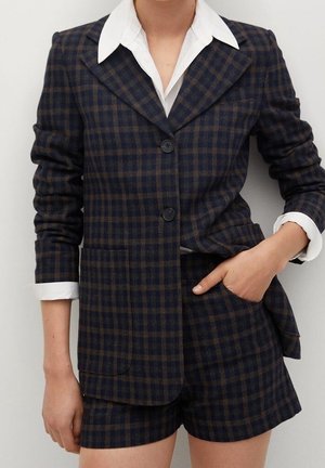 Femme portant un blazer à carreaux foncés et un short assorti avec une chemise blanche à col, une main dans la poche, sur un fond clair uni.