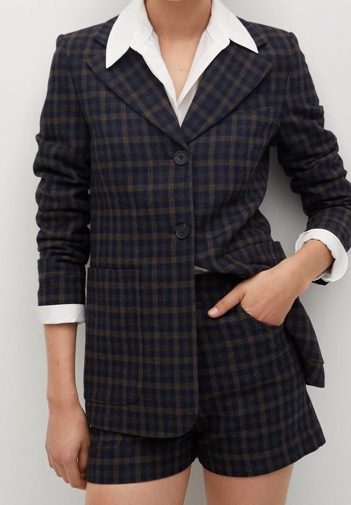 Femme portant un blazer à carreaux foncés et un short assorti avec une chemise blanche à col, une main dans la poche, sur un fond clair uni.