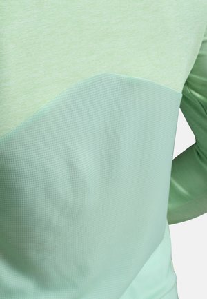 Nærbillede af en person iført en lysegrøn tekstureret bluse med lange ærmer og et gittermønster på den nederste halvdel.