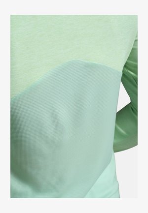 Nærbillede af en person iført en lysegrøn tekstureret bluse med lange ærmer og et gittermønster på den nederste halvdel.