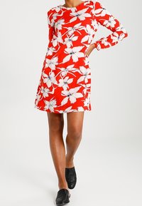 Robe fleurie rouge à manches longues avec des fleurs blanches, un col rond et une coupe décontractée, associée à des chaussures noires texturées.