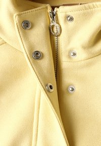 Veste jaune en tissu doux, dotée d'une fermeture éclair à l'avant, de pressions métalliques et d'un col rabattu. Texture lisse avec des coutures soignées.