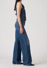 Femme debout en gilet en jean bleu et jean à larges jambes avec des chaussures noires pointues, vue de profil sur un fond uni.