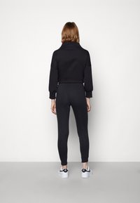 Juodas džemperis su aukštu apykakle ir numestais pečių siūlais, derinamas su juodomis legingo kelnėmis. Modelis vilki baltus sportinius batelius su juodais akcentais. Lygus paviršius.