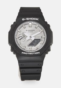 Montre-bracelet G-SHOCK avec un boîtier et un bracelet en résine noire, un cadran argenté avec des affichages analogiques et numériques, et un texte blanc pour les étiquettes de fonction.