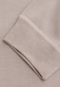 Beige tyg med en slät textur och fint stickat mönster. Vikta ärmslut som visar en kontrasterande ljusbeige sömdetalj.