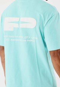 Camiseta de algodón azul claro de manga corta. Presenta un gran logo blanco y texto en la parte posterior, incluyendo "2015" y frases adicionales.