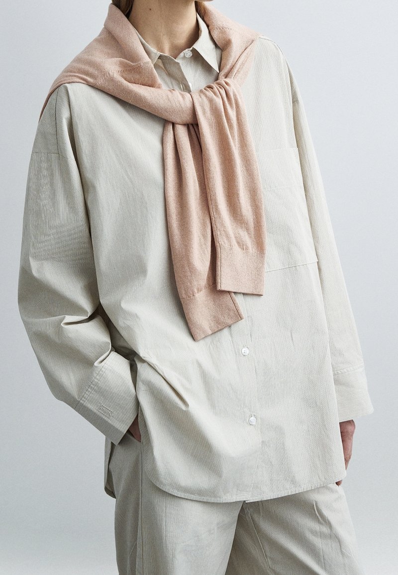 Personne portant une chemise et un pantalon en velours côtelé beige oversize avec un pull rose clair drapé sur les épaules et noué à l’avant.