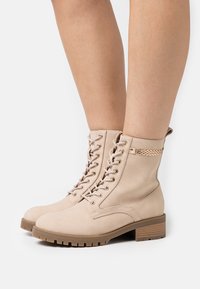 Bottines beiges à lacets fabriquées en cuir synthétique avec une surface texturée et un petit talon carré. Elles présentent des ferrures dorées et un accent de chaîne.