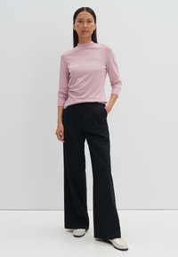 Helles rosa Langarmshirt mit hohem Kragen, kombiniert mit hochgeschnittenen, schwarzen weit geschnittenen Hosen und cremefarbenen Loafers. Schlichte, glatte Texturen.