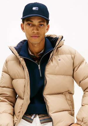 Beige pufferjack met capuchon, marineblauwe zip-up trui en donkere baseballpet. Kenmerkend door de zachte textuur en gelaagde look, met het Tommy Jeans-logo.