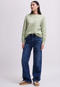 Ljusgrön långärmad topp med ett fiskbensmönster, som är parat med löst sittande mörkblå jeans och bruna slip-on skor med vita detaljer.