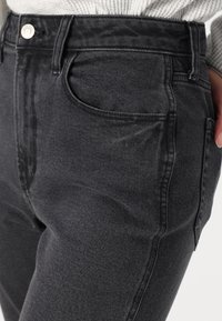 Jeans de ganga pretos com uma cintura alta, apresentando bolsos frontais, passantes para cinto e uma textura suave. O tecido tem pequenos detalhes de desbotamento.