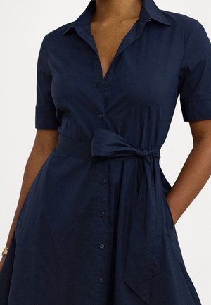 Kvinde iført en mørkeblå kjole med korte ærmer, knapper foran, bindebånd i taljen og en hånd i lommen.