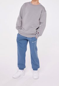 Felpa in cotone grigia con colletto rotondo e polsini a costine, abbinata a pantaloni tuta in pile blu con vita elasticizzata e logo. Sneakers bianche.