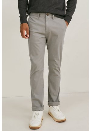 Mann mit grauen, hochgekrempelten Hosen, dunklem Pullover und weißen Turnschuhen mit braunen Sohlen, steht mit einer Hand in der Tasche.