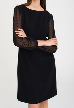 Vrouw die een zwarte jurk draagt met doorschijnende, lange opgeblazen mouwen met een gestippeld patroon, staand met gekruiste armen.