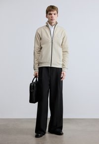 Ανοιχτό μπεζ ζακέτα με φερμουάρ και ψηλό κολάρο, με ριμπ μανσέτες και έμβλημα στο στήθος· συνδυασμένη με μαύρο παντελόνι φαρδιάς γραμμής και μαύρη τσάντα.