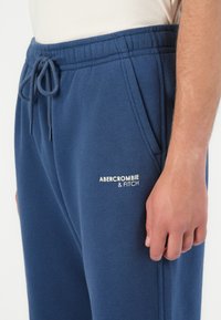 Zbliżenie na osobę w niebieskich dresach z elastycznym paskiem w talii, sznurkiem, bocznymi kieszeniami i logo "Abercrombie & Fitch".