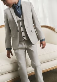 Garçon portant un costume trois pièces beige clair avec une chemise bleue et une cravate bleu marine, debout devant un canapé crème.