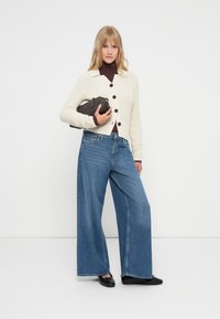 Cárdigan de punto crema con botones, cuello alto marrón, vaqueros de mezclilla anchos azules y zapatos negros. Sosteniendo un pequeño bolso de mano negro.