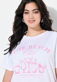 Weißes oversized T-Shirt mit einem pinken Grafikdesign von Cocktails und dem Text "THE BEACH Club." Kurze Ärmel und runder Ausschnitt mit pinken Nähten.