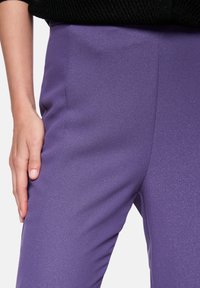 Purple, sparkly fabric trousers with a smooth texture, featuring a form-fitting design and side seam detail, held by a hand at the waist.
