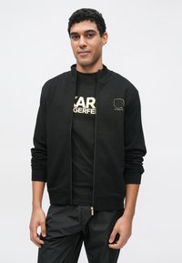 Μαύρο φούτερ με φερμουάρ, με ριμπ περιλαίμιο και μανσέτες, συνδυασμένο με μαύρο μπλουζάκι που έχει χρυσά γράμματα. Φοριέται με μαύρο παντελόνι.