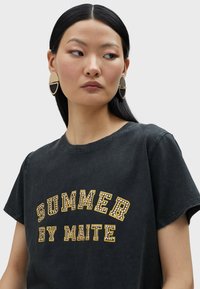 Camiseta de algodón negra con cuello redondo y mangas cortas, que presenta un gráfico de "SUMMER BY MAITE" en colores amarillo y blanco con tachuelas. Pendientes en dorado y negro.